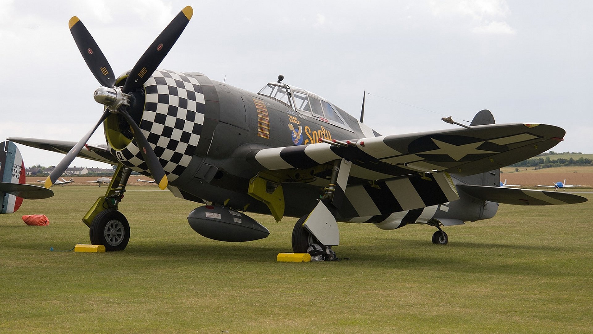 HD desktop wallpaper featuring a Republic P-47 Thunderbolt military aircraft with distinctive checkered nose and black-and-white wing stripes on a grassy airfield.