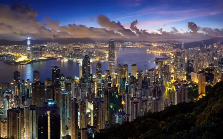A stunning HD wallpaper of Hong Kong's skyline at dusk, showcasing the vibrant city lights and skyscrapers reflecting on the water, surrounded by dramatic clouds.