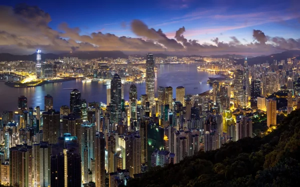 A stunning HD wallpaper of Hong Kong's skyline at dusk, showcasing the vibrant city lights and skyscrapers reflecting on the water, surrounded by dramatic clouds.