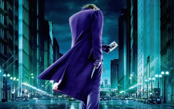 HD desktop wallpaper featuring the Joker from the movie The Dark Knight, standing in the middle of a city street at night, holding a knife and a Joker card, with buildings illuminated by streetlights.