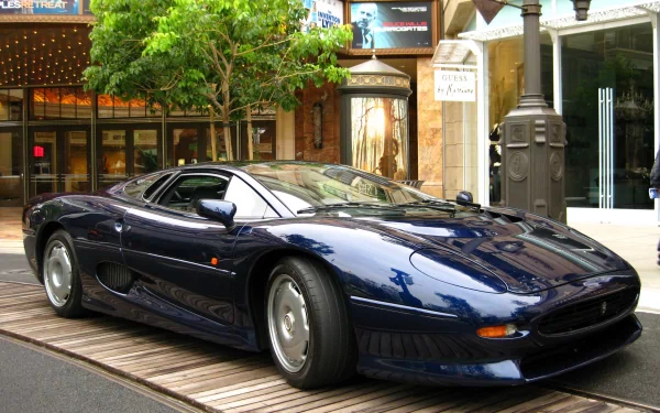 HD PC desktop wallpaper featuring a sleek, dark blue Jaguar XJ220 parked on a wooden platform in an urban setting with trees and storefronts.