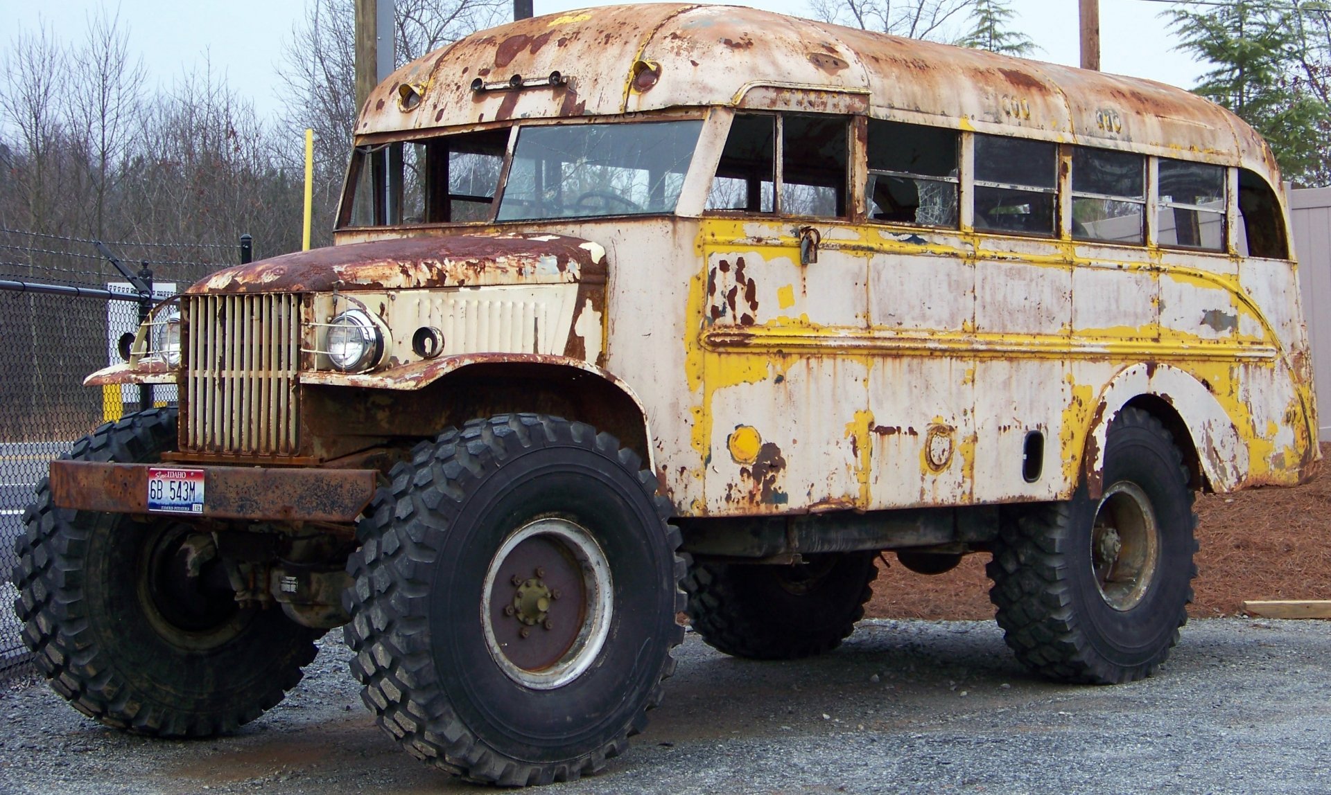 HD desktop wallpaper of a heavily rusted, vintage yellow bus with oversized off-road tires parked outdoors near a fence and trees.