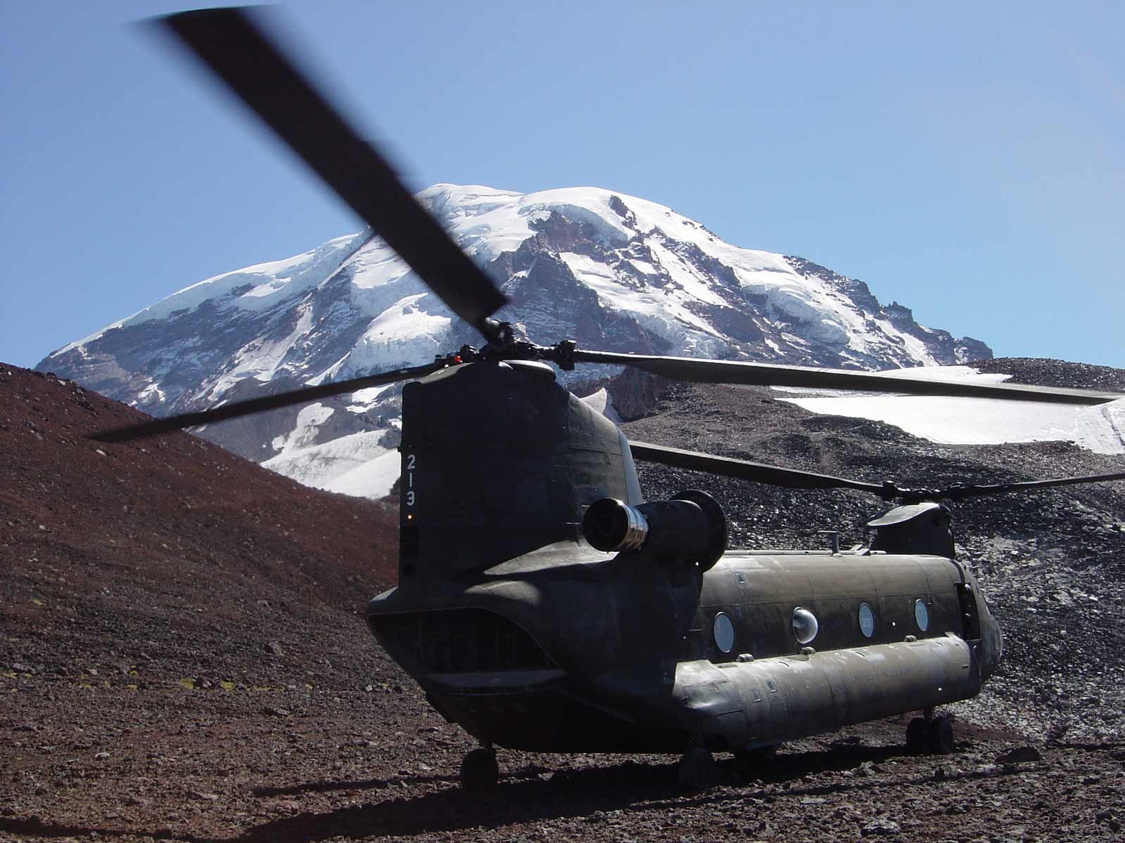 HD PC desktop wallpaper of a military Boeing CH-47 Chinook helicopter parked on rocky terrain with a snow-capped mountain in the background, rotor blades overhead.