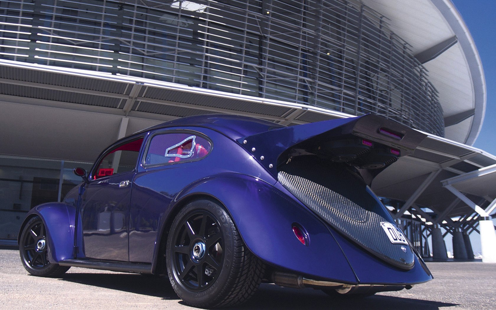 HD desktop wallpaper featuring a sleek, vintage Volkswagen vehicle in deep blue parked in front of a modern architectural structure.