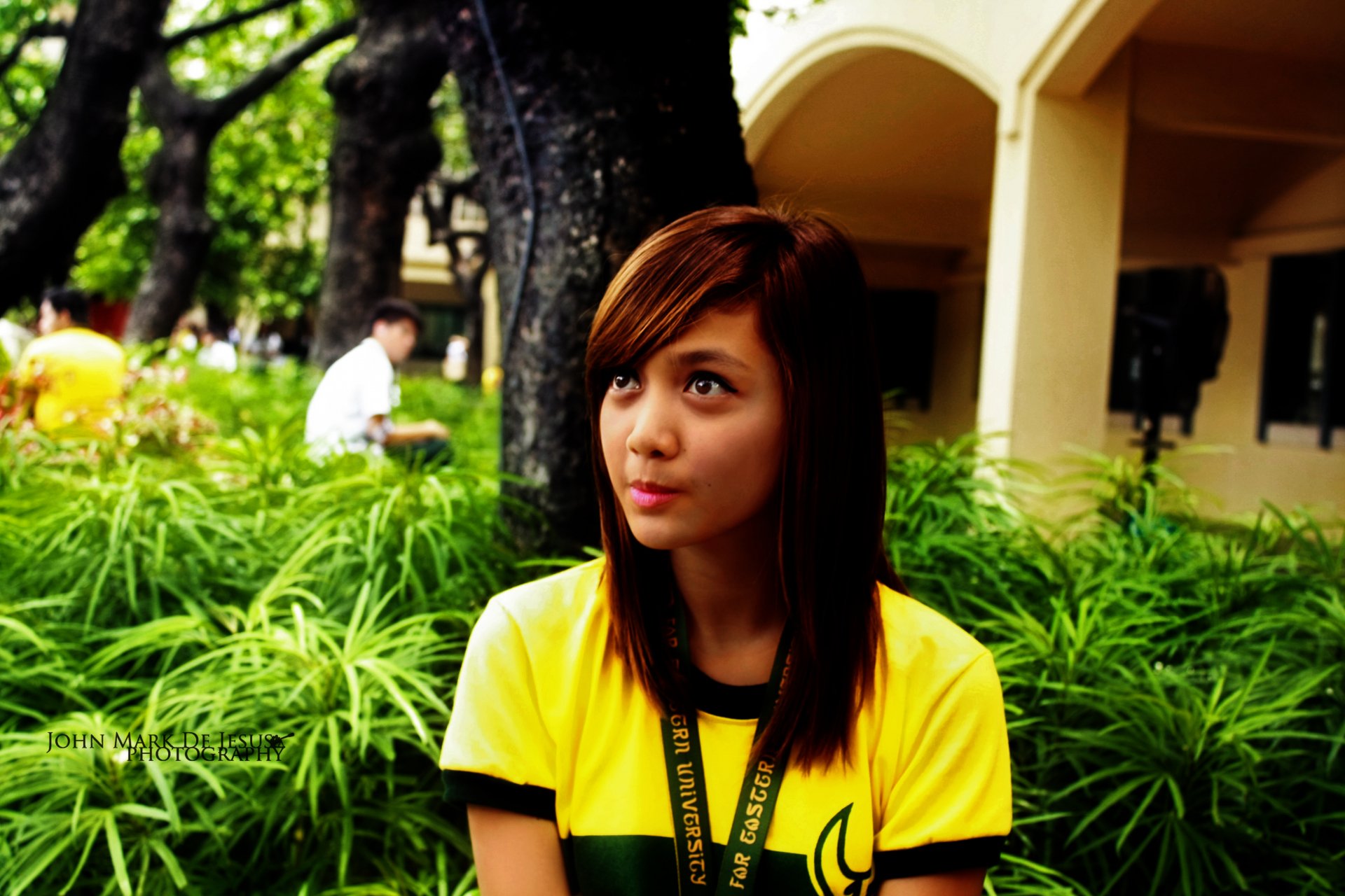 Pretty woman in a yellow shirt sitting among green foliage by a building, looking off-camera — 4K Ultra HD PC desktop wallpaper and background.