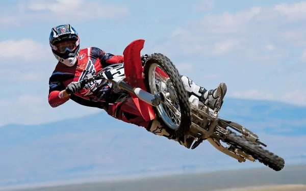 A motocross rider performs an impressive jump against a backdrop of blue skies and mountains, showcasing the thrill of the sport in this HD desktop wallpaper.