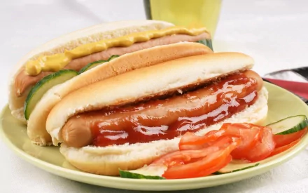 HD PC desktop wallpaper featuring a close-up of two hot dogs with mustard, ketchup, pickles, and tomato slices on a plate, highlighting classic American food.
