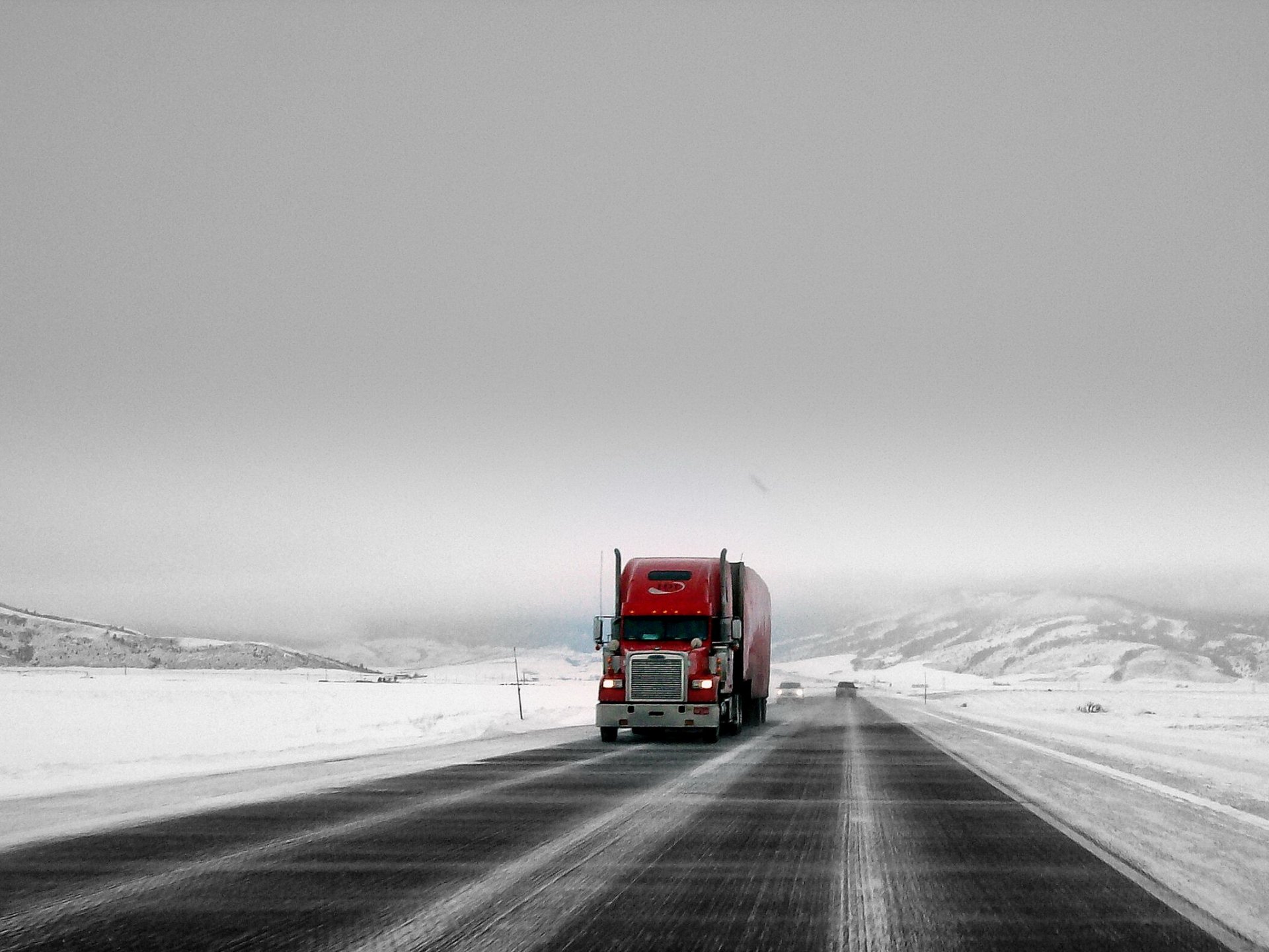 A red truck driving on a snowy, deserted highway under a cloudy gray sky, captured as an HD PC desktop wallpaper and background.