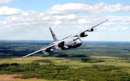 Military Lockheed C-130 Hercules aircraft flying over a lush green landscape under a partly cloudy sky, captured in high-definition for a desktop wallpaper background.