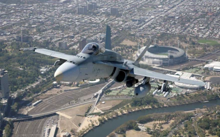 HD desktop wallpaper featuring a McDonnell Douglas F/A-18 Hornet military jet flying over a cityscape with a river and stadium visible below.