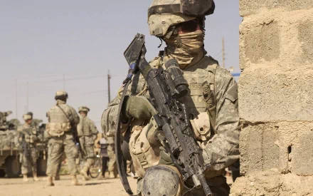 HD military PC desktop wallpaper showing a soldier in full combat gear holding a rifle beside a stone wall with other soldiers in the background.