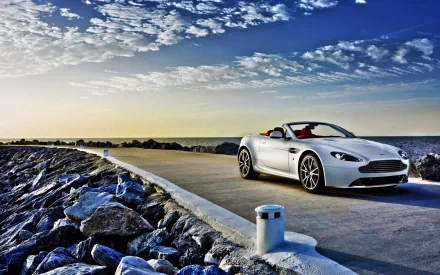 Silver Aston Martin V8 Vantage convertible driving on a coastal road under a vibrant blue sky, captured as an HD PC desktop wallpaper and background.