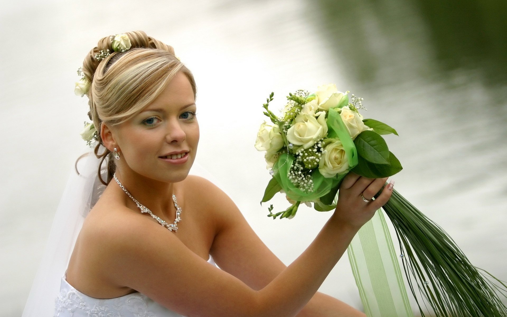 HD desktop wallpaper of a smiling bride holding a bouquet of white roses, posing outdoors by a calm water background.