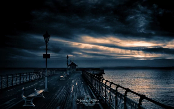 HD desktop wallpaper of a man-made pier at sunset with a bench along the wooden walkway, calm sea, and dramatic clouds overhead.