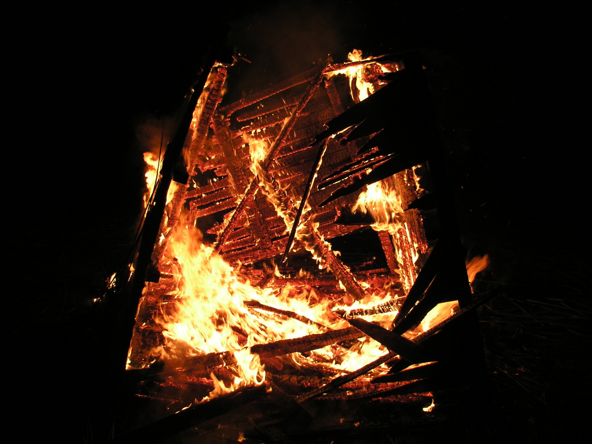 A high-definition desktop wallpaper featuring a vibrant photograph of a burning fire, showcasing intense flames that consume a wooden structure against a dark background.