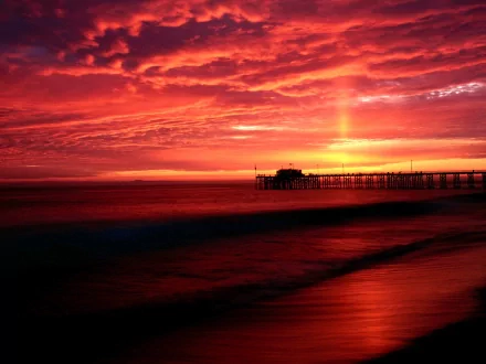 A stunning HD desktop wallpaper featuring a vibrant red sunset over a pier, with swirling clouds reflecting the rich hues of the sky on the calm water.