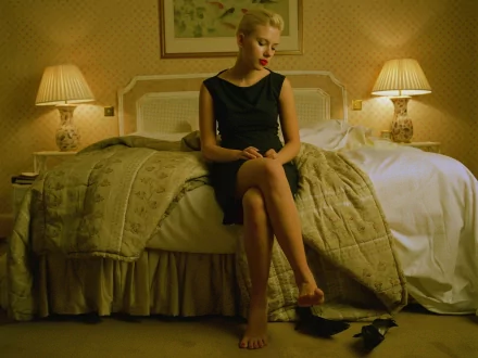 HD wallpaper depicting Scarlett Johansson seated at the edge of a bed in a warmly lit, elegantly decorated bedroom. She is wearing a black dress, looking down thoughtfully.