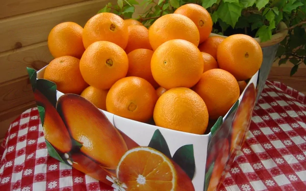 HD desktop wallpaper featuring a box filled with fresh, vibrant oranges on a red and white checkered tablecloth. The background shows green foliage adding a touch of natural beauty.