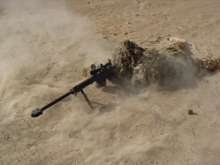 HD desktop wallpaper showing a camouflaged military sniper lying in a dusty environment, gripping a long-range rifle, encompassed by sand and haze. Tags: weapon, soldier, military, sniper.
