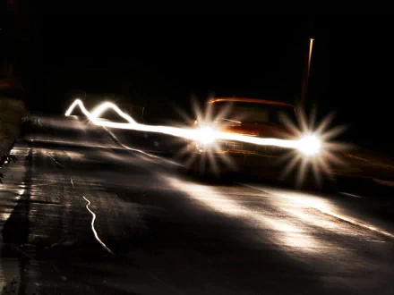 HD desktop wallpaper featuring a car driving at night with bright headlights illuminating a dark road, creating dynamic light trails and reflections.