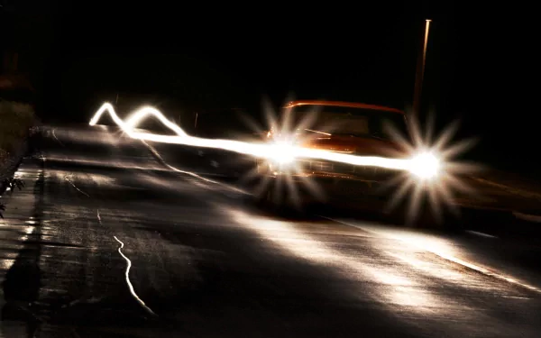 HD desktop wallpaper featuring a car driving at night with bright headlights illuminating a dark road, creating dynamic light trails and reflections.