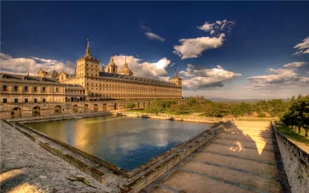 HD desktop wallpaper featuring the majestic El Escorial, a grand historical complex in Spain, beautifully bathed in sunlight under a vivid blue sky with clouds reflected in a serene pond.