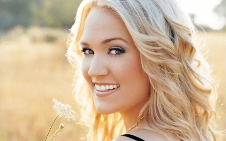 A close-up of a smiling woman with long, wavy blonde hair in a sunlit field, holding a small flower. This HD wallpaper highlights the beauty of music artist Carrie Underwood.