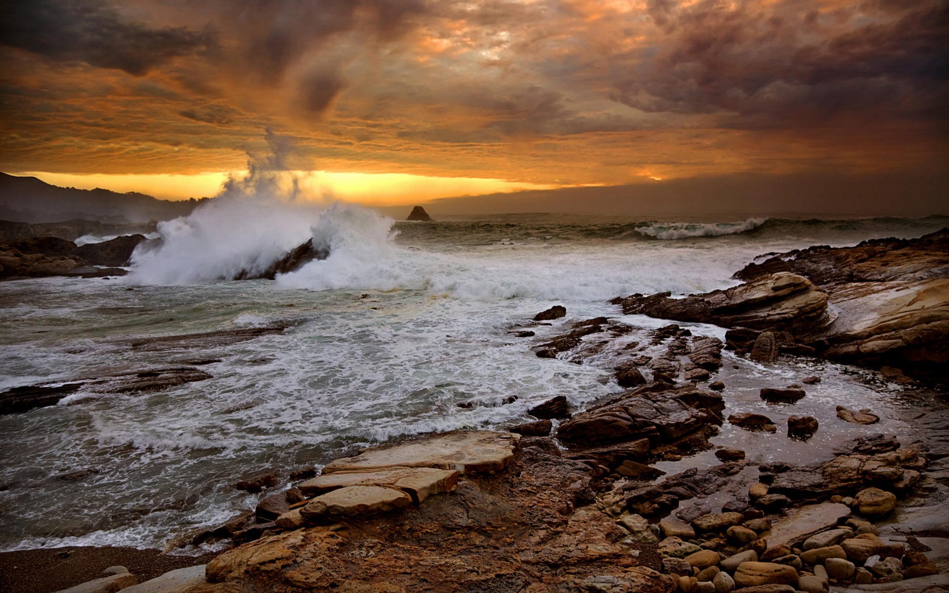 A stunning sunset casts golden hues over a turbulent ocean, with waves crashing against rocky shores. The vibrant clouds and horizon create a breathtaking natural scene.