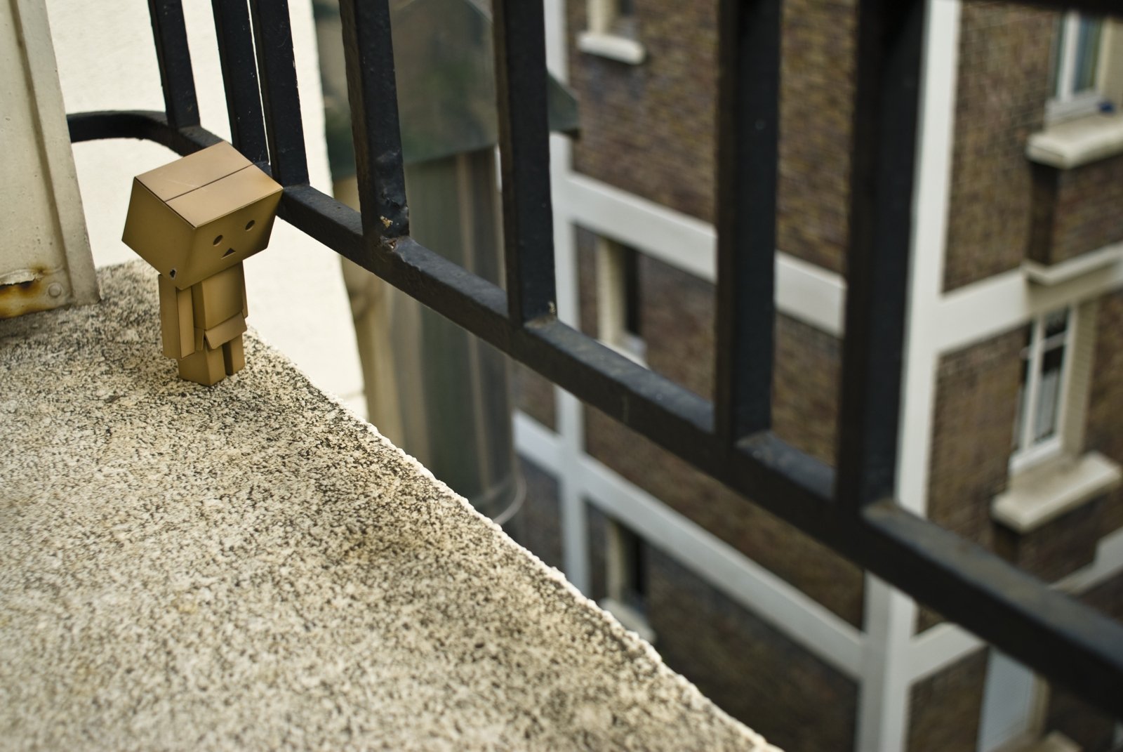 HD PC desktop wallpaper featuring a small cardboard Danbo figure standing on a balcony ledge, overlooking an urban building exterior.