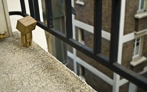 HD PC desktop wallpaper featuring a small cardboard Danbo figure standing on a balcony ledge, overlooking an urban building exterior.