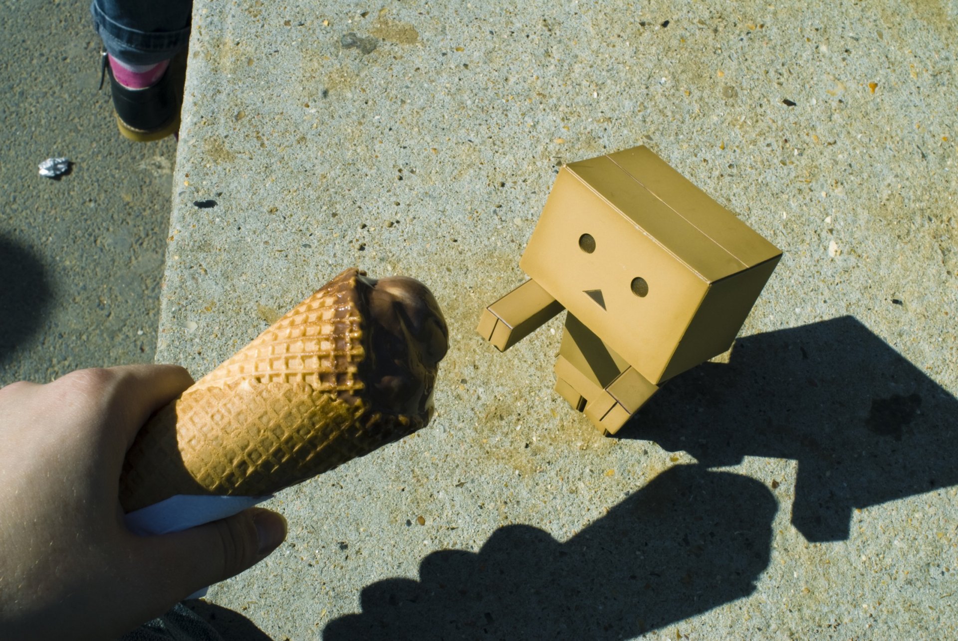 HD PC desktop wallpaper featuring a small cardboard Danbo figure curiously looking at a hand holding a chocolate ice cream cone on a concrete surface.