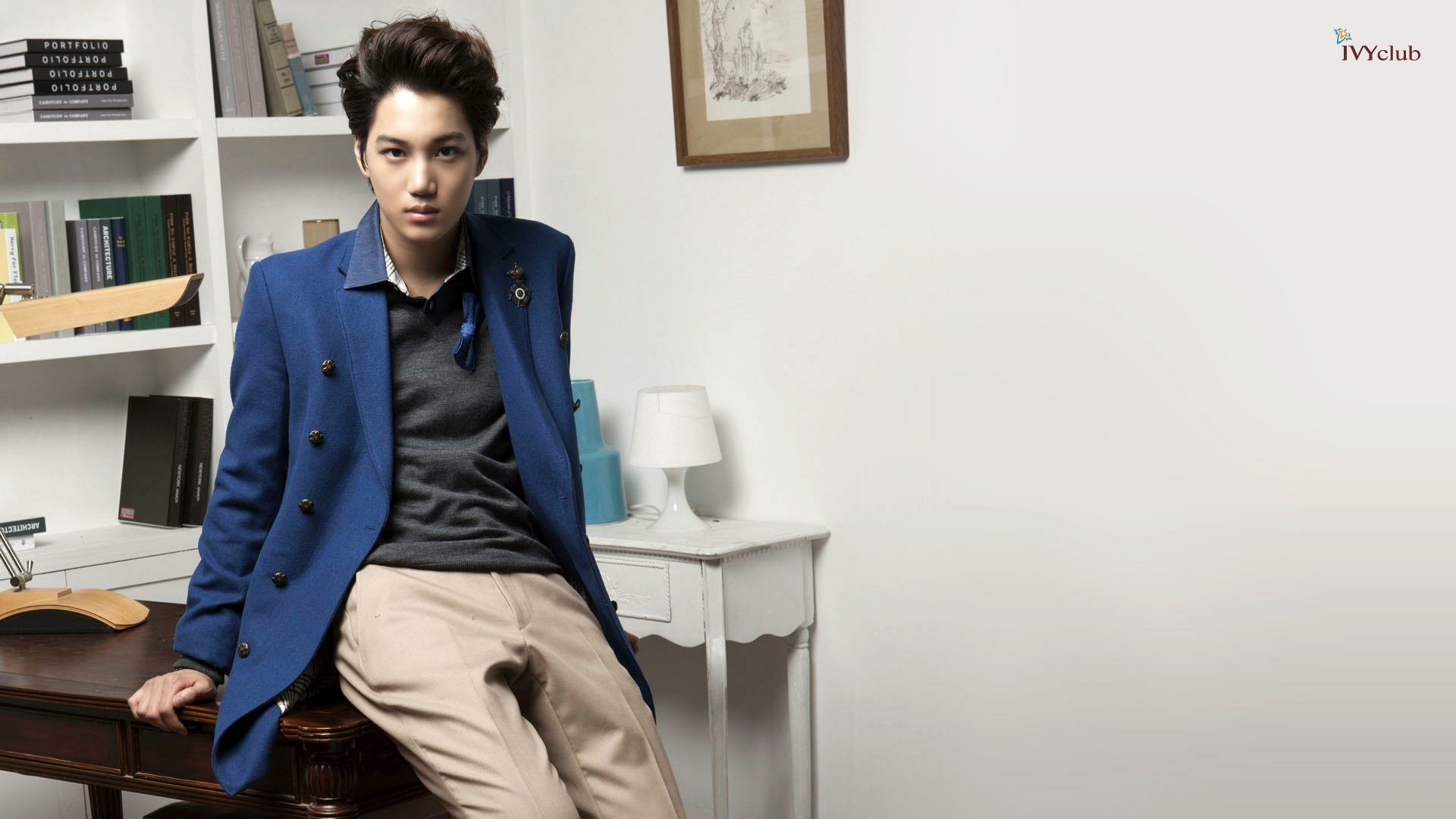A stylish HD wallpaper featuring a member of EXO seated at a desk, dressed in a blue blazer with a contemporary background of shelves and decor elements.