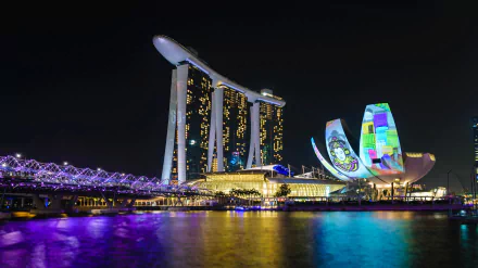 Night view of Singapore's Helix Bridge and Marina Bay Sands, illuminated against the dark sky, showcasing modern man-made architecture reflected on the water.