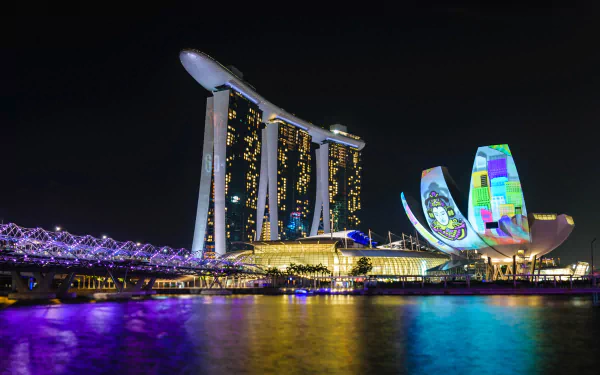 Night view of Singapore's Helix Bridge and Marina Bay Sands, illuminated against the dark sky, showcasing modern man-made architecture reflected on the water.
