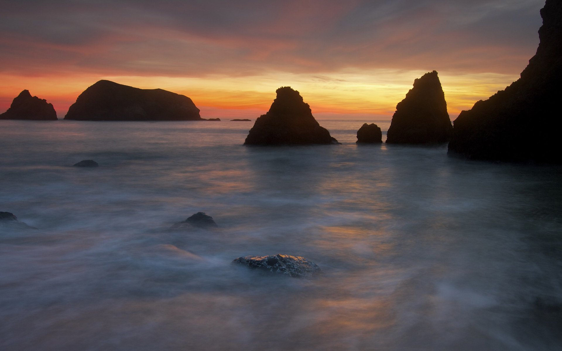 2K Quad HD PC desktop wallpaper/background — nature seascape at sunset: silhouetted sea stacks, smooth misty waves and a glowing orange‑purple sky.