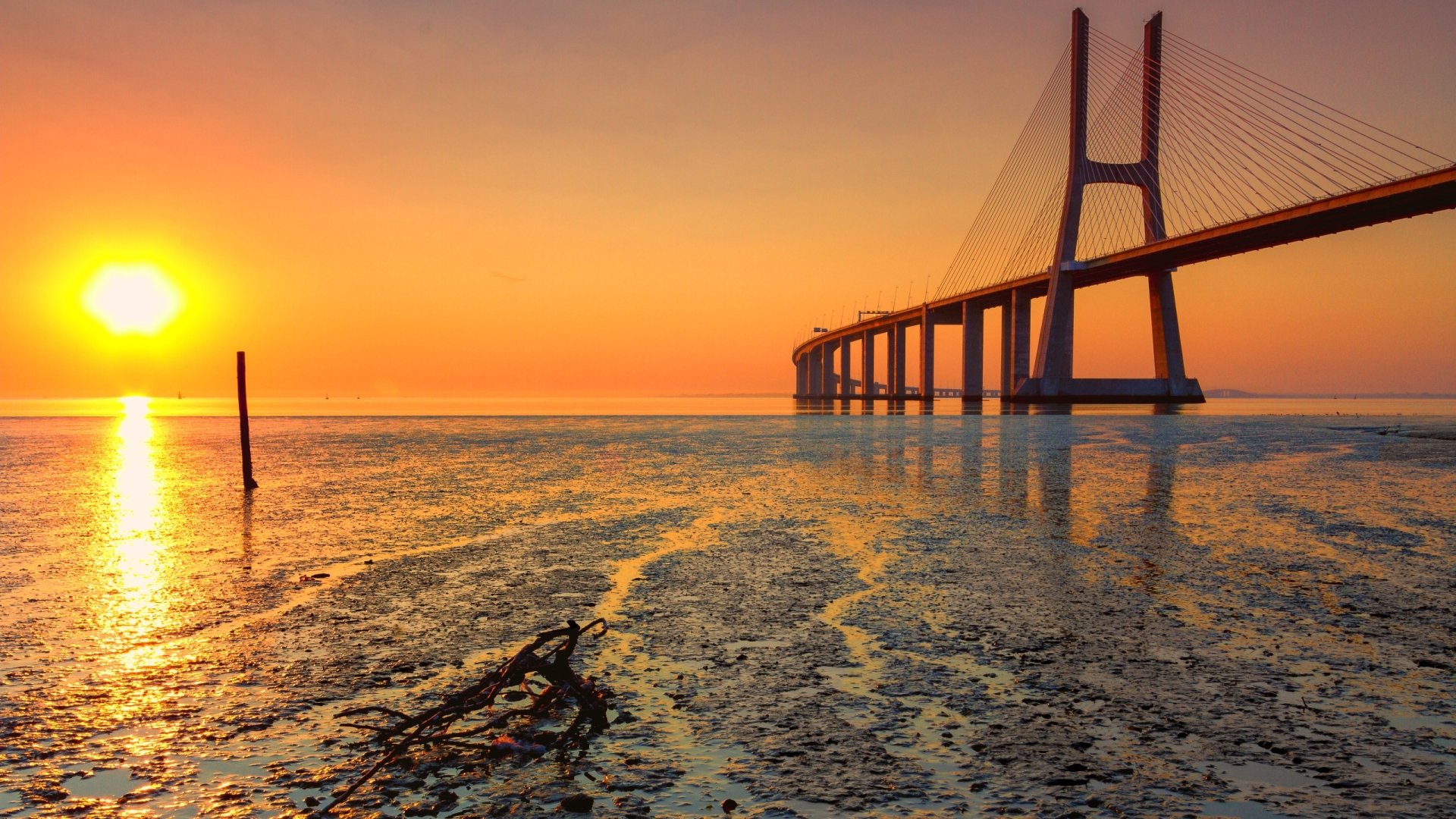 Vasco da Gama Bridge at golden sunset over tidal flats, vivid orange sky and reflective water, a man-made landmark presented as a 2K Quad HD PC desktop wallpaper and background.