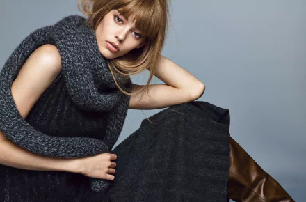 HD desktop wallpaper of a woman with light brown hair wearing a chunky gray scarf and black outfit against a plain gray background.