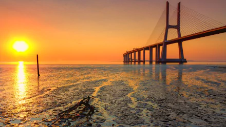 Vasco da Gama Bridge at golden sunset over tidal flats, vivid orange sky and reflective water, a man-made landmark presented as a 2K Quad HD PC desktop wallpaper and background.