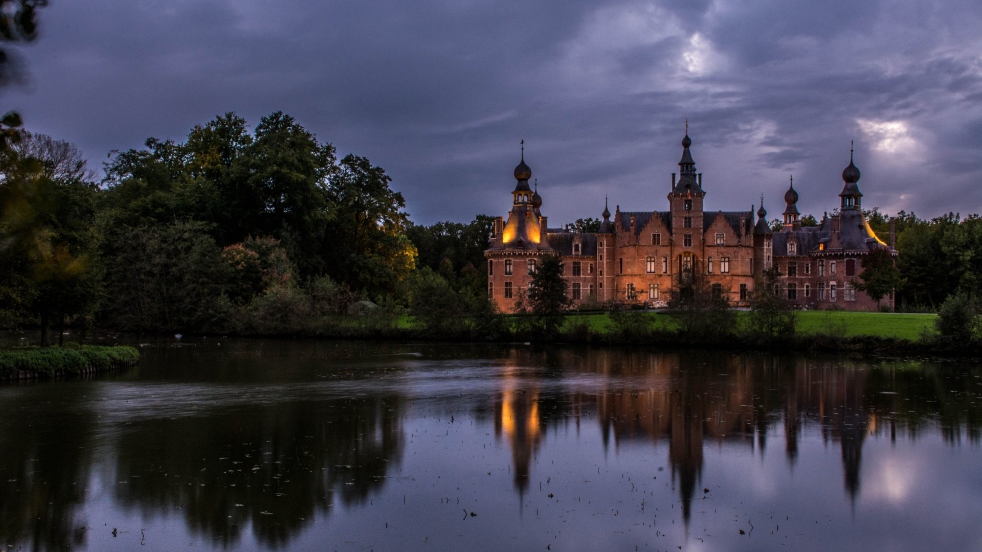 Ooidonk Castle at dusk, glowing turrets reflected in a tranquil moat beneath brooding clouds — 2K Quad HD PC desktop wallpaper of a man-made historic castle.