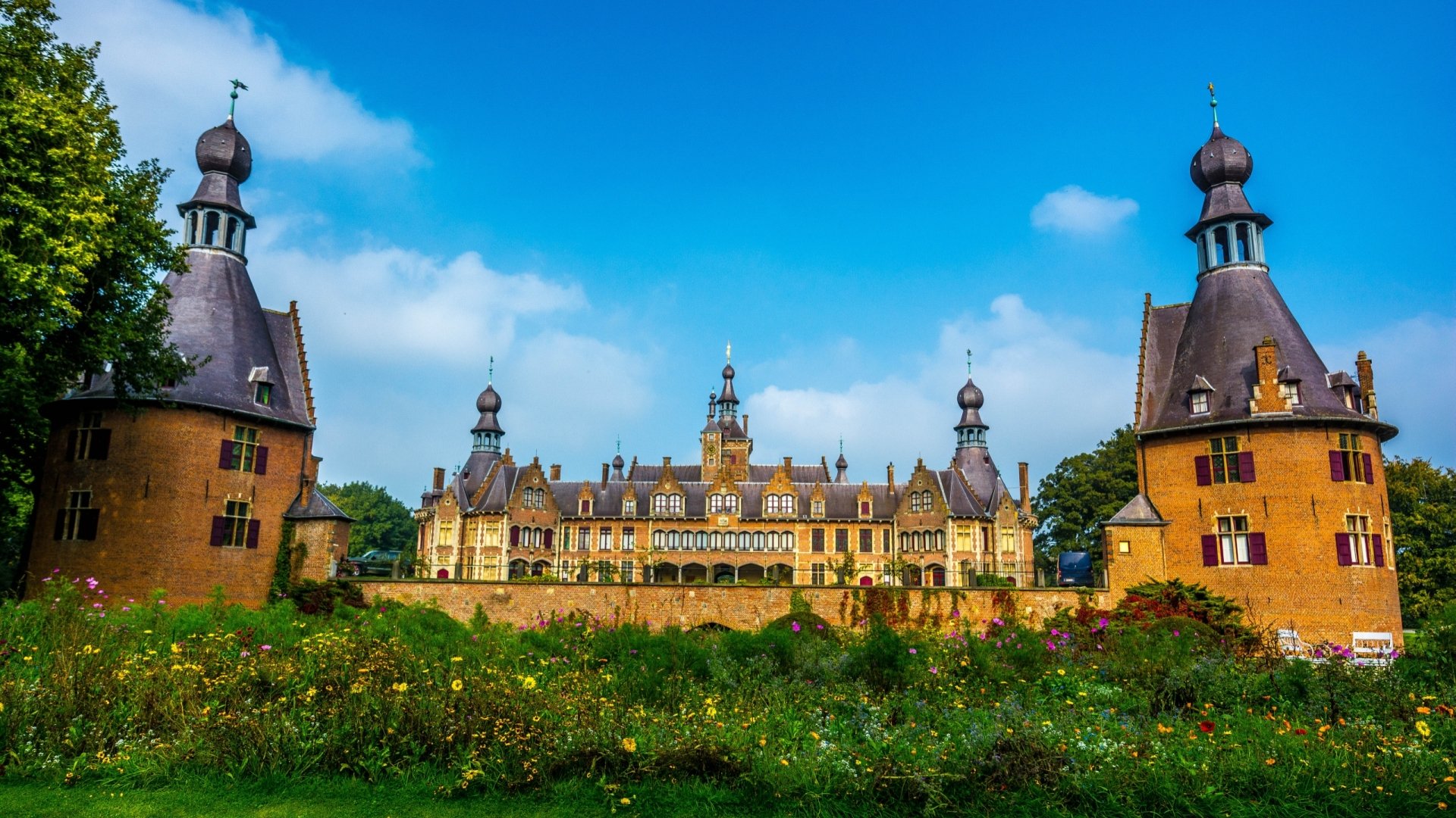 HD desktop wallpaper showcasing the man-made Ooidonk Castle under a bright blue sky, framed by lush greenery and vibrant flowers in the foreground.