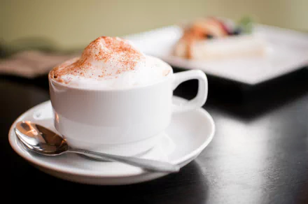A close-up of a frothy cappuccino in a white cup sits on a saucer with a spoon, accompanied by a plate of pastries, showcased in a 4K Ultra HD background.
