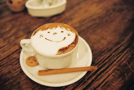A cozy cappuccino with a smiling foam design sits on a wooden table, accompanied by a cinnamon stick and a cookie, creating a warm and inviting atmosphere. HD desktop wallpaper.