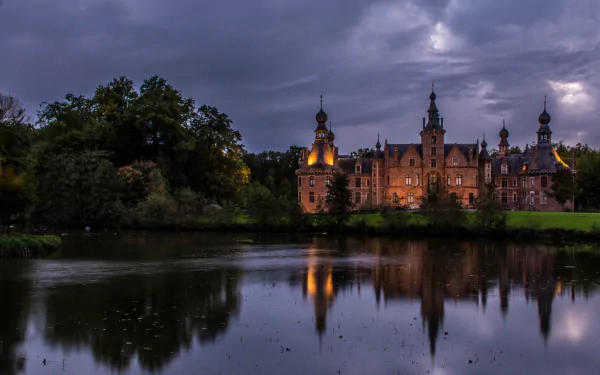 Ooidonk Castle at dusk, glowing turrets reflected in a tranquil moat beneath brooding clouds — 2K Quad HD PC desktop wallpaper of a man-made historic castle.