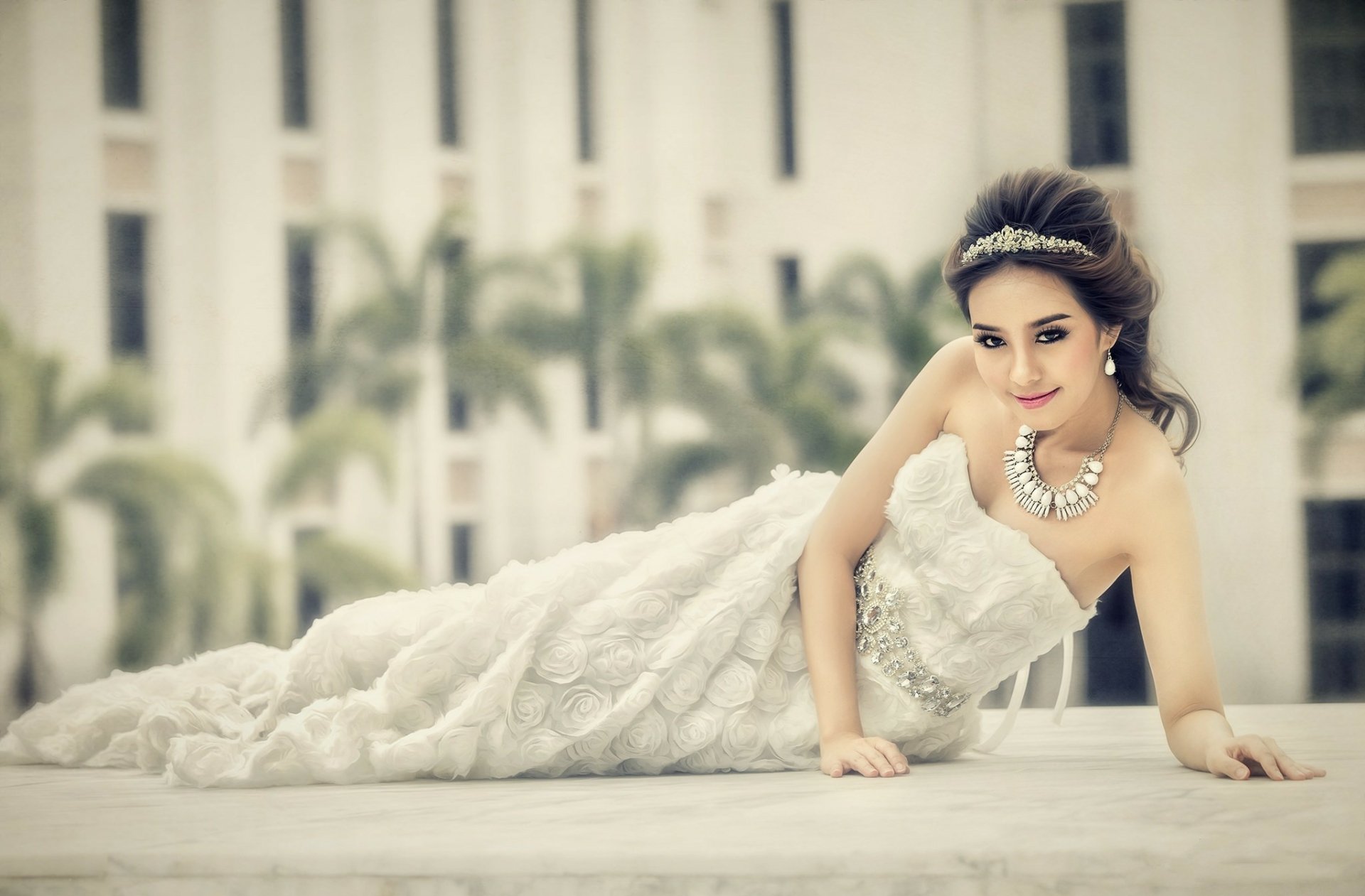 HD desktop wallpaper featuring an Asian woman model in a white dress adorned with a statement necklace, set against a blurred urban backdrop with palm trees.
