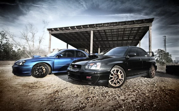 HD PC desktop wallpaper featuring two Subaru Impreza vehicles parked on a dirt surface under a rustic metal-roofed shelter against a cloudy sky.