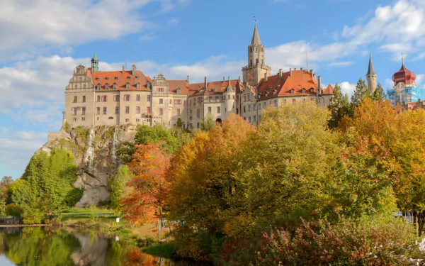 Man-made Sigmaringen Castle perched on a limestone cliff above a reflective river, framed by autumn trees and a blue sky — HD PC desktop wallpaper and background.