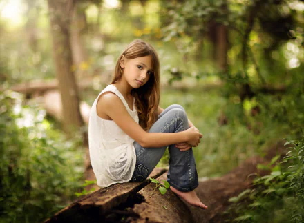 A barefoot little girl with long hair, wearing a white shirt and jeans sits on a log in a serene, bokeh-filled forest scene, evoking a calm and natural mood. This is an HD desktop wallpaper and background.