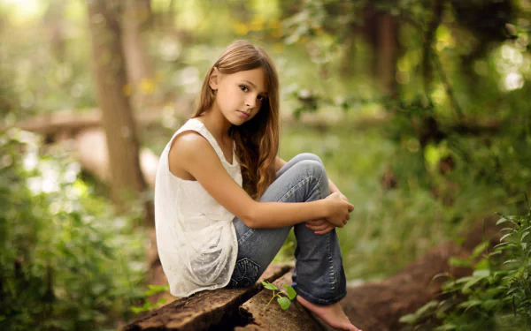 A barefoot little girl with long hair, wearing a white shirt and jeans sits on a log in a serene, bokeh-filled forest scene, evoking a calm and natural mood. This is an HD desktop wallpaper and background.