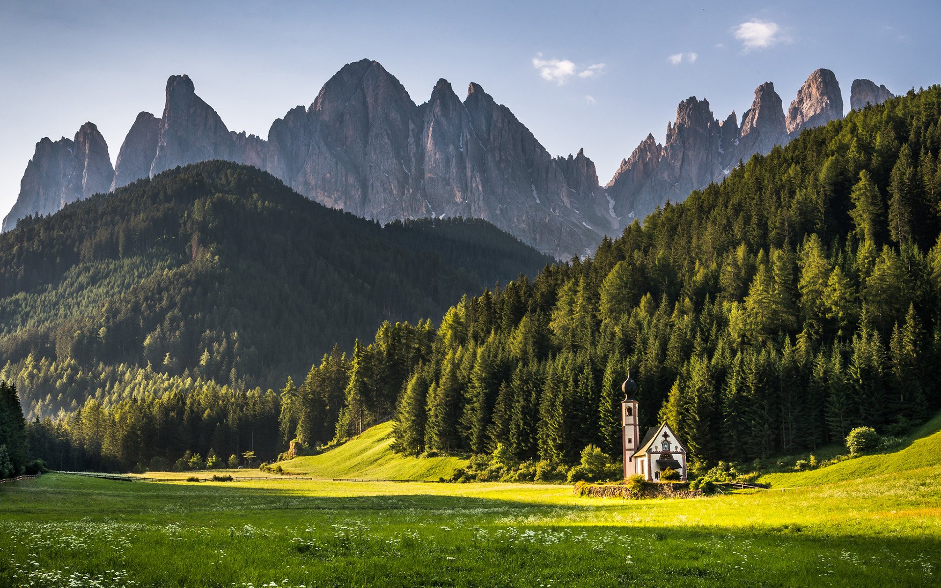 HD landscape photography of a serene mountain range with sharp peaks, dense forest, and a small church in a sunlit green meadow, captured for PC desktop wallpaper.