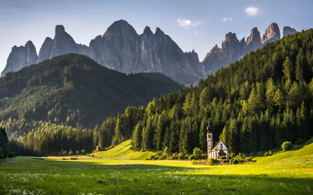 HD landscape photography of a serene mountain range with sharp peaks, dense forest, and a small church in a sunlit green meadow, captured for PC desktop wallpaper.